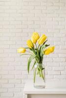 gelbe Tulpen in einer Glasvase auf einem weißen Backsteinmauerhintergrund