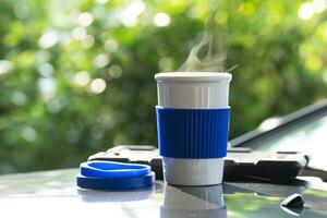 heiß Keramik Weiß Kaffee Tasse mit Rauch platziert draußen das Auto Instrumententafel beim Sonnenaufgang im das Morgen, im ein natürlich Hintergrund. heiß Kaffee trinken Konzept, selektiv Fokus, Sanft Fokus. foto