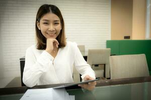 schön asiatisch Geschäft Frau im Nahansicht Schuss ist Vertrauen im ihr besitzen Der Umsatz und Sitzung während halten Tablette mit Lächeln auf ihr Gesicht, Büro Mädchen Konzept foto