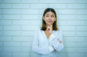 schön asiatisch Geschäft Frau ist Stehen im ein nachdenklich Pose beim das Kopieren Raum mit ein Weiß Backstein Hintergrund, Digital Marketing. foto