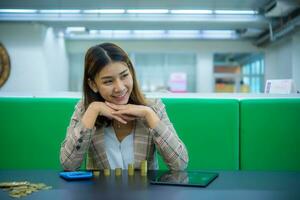 jung schön asiatisch Geschäft Frau ist Sitzung während suchen beim das Kopieren Raum mit ein Lächeln Dort sind Haufen von Gold Münzen Tablets, und Taschenrechner platziert auf ein schwarz Tisch, Digital Marketing. foto