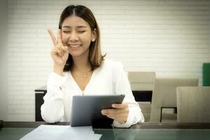 schön asiatisch Geschäft Frau ist ermutigen du selber zu Arbeit und Sitzung zeigen Sieg Zeichen auf ihr Wange während halten Tablette mit Augen schließen. foto