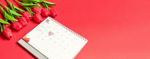 das Konzept zum Valentinstag Tag auf 14 .. Februar 2022. Nahaufnahme, selektiv Fokus, verschwommen Hintergrund, das Kennzeichen Herz auf 14 .. Kalender von Februar mit rot Tulpen Blume auf das rot Hintergrund auf ein Schreibtisch foto