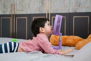 asiatisch Junge Lügen auf das Matratze auf ein heiß Tag spielen mit ein tragbar Ventilator glücklich foto