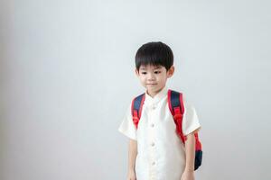 asiatisch Junge tragen traditionell thailändisch Kleidung, Stehen mit ein Schule Tasche. vorbereiten zu gehen zu Schule auf ein Weiß Hintergrund foto