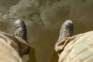 Herren Beine im tarnen Hose und alt Stiefel baumeln über das Wasser. foto