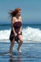 glücklich und begeistert Frau im lange Kleid Laufen durch Wasser von brechen Ozean Wellen auf Strand foto