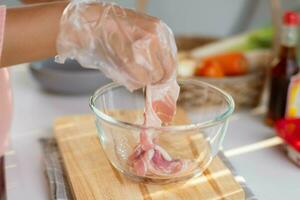 weiblich Hände im ein Plastik Handschuh Picks oben ein geschnitten Schweinefleisch Bauch in ein Schüssel. Frau vorbereiten Essen im das Küche beim heim. foto