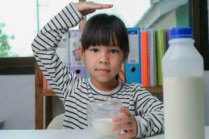 süß asiatisch Mädchen Trinken ein Glas von Milch beim Zuhause im Leben Zimmer. wenig Mädchen Trinken Milch im das Morgen Vor gehen zu Schule. gesund Essen im Kindheit. foto