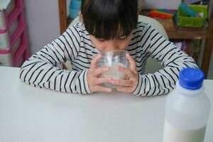 süß asiatisch Mädchen Trinken ein Glas von Milch beim Zuhause im Leben Zimmer. wenig Mädchen Trinken Milch im das Morgen Vor gehen zu Schule. gesund Essen im Kindheit. foto