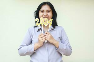 glücklich asiatisch jung Frau Büro Arbeiter schreiend zu feiern Neu Jahre Vorabend halten Gold und Weiß Kerzen 2024 Zahlen und erziehen Hand foto