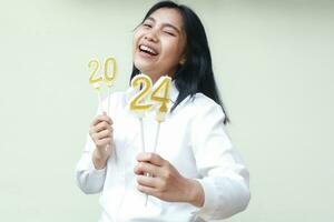 aufgeregt asiatisch jung Frau Mitarbeiter Lachen zu Kamera und Show 2024 Zahl Kerzen angehoben auf ihr Hand tragen formal Weiß Shirt, isoliert, Neu Jahre Vorabend Konzept foto