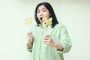 asiatisch jung Frau überrascht mit öffnen Mund tragen Grün Über Größe Hemd mit halten 2024 Nummer Kerze, suchen zu Kamera, isoliert Über Weiß Hintergrund foto