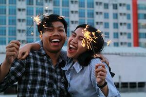 aufgeregt asiatisch jung Paar halten Verbrennung Wunderkerze und Lachen aussehen beim Kamera zu feiern Neu Jahre Vorabend mit städtisch Gebäude Hintergrund, Hipster Teenager tragen beiläufig Hemd foto