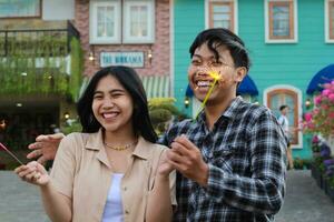 Lachen asiatisch Paar Teen lächelnd zu Kamera mit halten Wunderkerzen im das bunt Haus Hof. Hipster freunde feiern Neu Jahr Vorabend foto