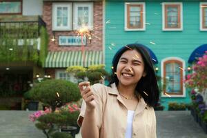 Porträt von schön glücklich jung asiatisch Frau halten Wunderkerzen Feuerwerk zu feiern Neu Jahr Vorabend mit Garten Party Stehen im draussen Jahrgang Haus Hof foto