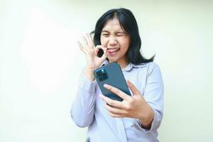 aufgeregt asiatisch jung Geschäft Frau halten Zelle Telefon mit leeren Bildschirm mit in Ordnung Zeichen Geste, zeigen imaginär Produkt, lächelnd weiblich aussehen Kamera tragen formal Hemd isoliert im Weiß Hintergrund foto