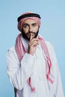 Muslim Mann tragen traditionell Kleider zeigen ruhig Geste und berühren Finger zu Lippen Porträt. arabisch Person Herstellung tsss Zeichen und suchen beim Kamera mit ernst Ausdruck foto