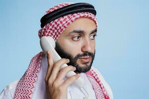 Muslim Mann tragen traditionell kariert Schal Sprechen auf Festnetz Telefon Nahaufnahme. arabisch Person im Kopfschmuck mit Seil Band antworten retro Telefon Anruf mit neutral Gesicht Ausdruck foto