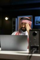detailliert Aussicht von müde Mann im arabisch Kleidung Sitzung beim Büro Schreibtisch und mit Laptop zu Verhalten Internet Forschung. Muslim Mann mit Minicomputer auf das Tisch, suchen müde und Leiden von Kopfschmerzen. foto