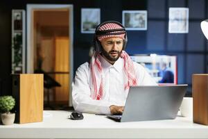 ein traditionell Mitte östlichen Unternehmer fleißig funktioniert im ein modern Büro, nutzen kabellos Technologie zum Kommunikation und Forschung. jung arabisch Mann mit seine kabellos Kopfhörer und Laptop. foto
