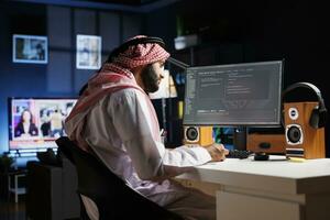 gewidmet Software Programmierer tragen traditionell Arabisch Kleidung ist sitzend beim seine Zuhause Büro Schreibtisch Prüfung Datenbank Code auf Computer. Muslim Mann Überprüfung das Programmierung Sprache auf das pc Monitor. foto