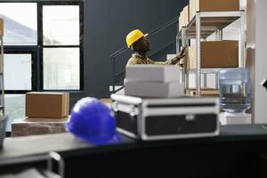 afrikanisch amerikanisch Post Büro Arbeiter suchen Kunde Paket im Lager Zimmer Regal. Versand Manager im schützend Helm finden Karton Box mit Klient bestellen im Post- Lagerhaus foto