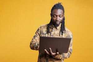 afrikanisch amerikanisch Mann halten Laptop Computer Surfen Webseiten auf Internet, Stehen im Studio Über Gelb Hintergrund. jung Erwachsene mit tragbar Gerät zu navigieren auf Sozial Medien foto