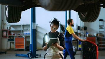 qualifiziert Techniker Stehen unterhalb suspendiert Auto im Garage Arbeitsplatz, mit Arbeit Licht zu prüfen zum Schäden und Tablette zu bestellen Neu ersetzen Teile nach finden Probleme während Routine Untersuchung foto