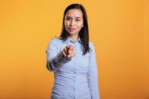 glücklich aufgeregt Filipino Frau zeigen Index Finger beim Kamera, posieren im Studio Über Gelb Hintergrund. froh lächelnd jung Erwachsene Person zeigen Zeigefinger Werbung Werbung Produkt foto