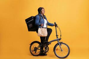 afrikanisch amerikanisch Lieferung Frau halten Papier Tasche während liefern Mittagessen bestellen zu Klient mit Fahrrad wie Transport. Restaurant Kurier Tragen Mitnahme Thermal- Rucksack, wegbringen Bedienung Konzept foto