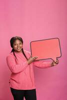 lächelnd afrikanisch amerikanisch zuversichtlich Frau halten rot Karton Rede Blase Stehen im Studio auf Rosa Hintergrund. froh jung Erwachsene halten leer Vorlage Ankündigung Plakat. foto