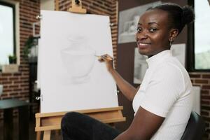 glücklich inspiriert jung afrikanisch amerikanisch Frau Erstellen einzigartig Stück von Kunst während kreativ Kunst Werkstatt, Sitzung beim Staffelei und lächelnd beim Kamera, genießen Zeichnung Lektion, Entwicklung ihr besitzen Kreativität foto