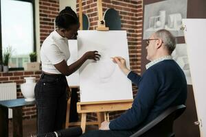 vielfältig anders alt Kunst Schule Studenten kommunizieren während Zeichnung Lektion, suchen beim Kunstwerk auf Segeltuch und diskutieren Skizzierung Techniken. afrikanisch amerikanisch Mädchen Portion Senior Mann beim Kunst Klasse foto