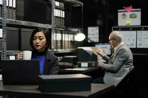 Buchhaltung Angestellte im Beratung bürokratisch Arbeitsplatz gefüllt mit Geschäft Ordner. asiatisch Frau genießen Tasse von Kaffee und Senior Mitarbeiter Arbeiten im administrative Datei Zimmer Büro foto