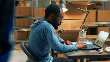 afrikanisch amerikanisch Mann Analysieren Lager und Aufträge, tun Inventar auf Laptop beim Warenhaus Raum. männlich Mitarbeiter mit Logistik und finanziell Planung zu senden Fan-Shop zum Geschäft planen. foto