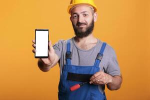 Konstruktion Arbeiter halten Zelle Telefon mit leeren Bildschirm zum Werbung im Vorderseite von Kamera. Fachmann Baumeister mit schwer Hut gegen Gelb Hintergrund im Studio Schuss. foto