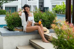 glücklich jung schön Bräunen Frau im stilvoll Boho Weiß Strandkleidung Sitzung in der Nähe von tropisch Schwimmbad im Luxus Hotel und genießen Orange Cocktail oder Saft. Strand Party. foto