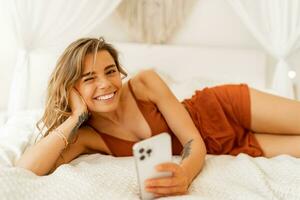 froh heiter Mädchen mit Smartphone während ruhen im Schlafzimmer. warm Farben, gemütlich Zuhause Stimmung. tragen Pyjama. foto