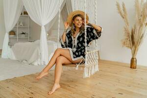 gemütlich Zuhause Atmosphäre. schön stilvoll blond Frau im Boho Kleid posieren im stilvoll Schlafzimmer. Dame Sitzung auf hängend schwingen . foto