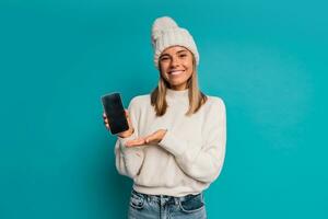 Studio Winter Porträt von lächelnd blond Frau im Weiß Hut und Whool Sweatshirt ein halten mobyle Telefon ,posieren im Studio Über türkis Hintergrund. foto