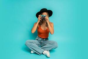 schön blond Frau im stilvoll Sommer- Outfit halten retro Kamera , Sitzung auf Fußboden im Studio auf Blau Hintergrund. Ferien Stimmung. foto