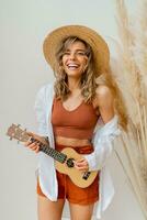 lächelnd anmutig Frau im Sommer- Outfit mit Stroh Hut spielen Ukulele Gitarre Über Weiß Hintergrund im Studio mit Pampas Gras Dekor. foto