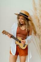 lächelnd anmutig Frau im Sommer- Outfit mit Stroh Hut spielen Ukulele Gitarre Über Weiß Hintergrund im Studio mit Pampas Gras Dekor. foto