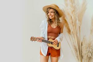 lächelnd anmutig Frau im Sommer- Outfit mit Stroh Hut spielen Ukulele Gitarre Über Weiß Hintergrund im Studio mit Pampas Gras Dekor. foto