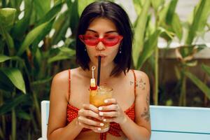 atemberaubend bräunen Frau im retro Sonnenbrille entspannend im tropisch Strand Verein im stilvoll rot oben und Orange kurze Hose. Trinken lecker Limonade. Ferien und Feiertage. foto