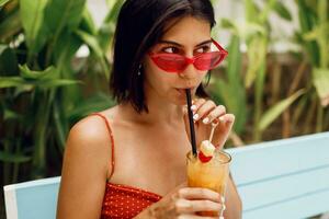 atemberaubend bräunen Frau im retro Sonnenbrille entspannend im tropisch Strand Verein im stilvoll rot oben und Orange kurze Hose. Trinken lecker Limonade. Ferien und Feiertage. foto