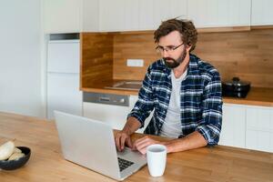 gut aussehend Mann Freiberufler mit Laptop studieren online Arbeiten von heim, glücklich beiläufig Kerl Tippen auf Notizbuch Surfen Internet suchen beim Bildschirm genießen entfernt Job sitzen beim Tisch. foto