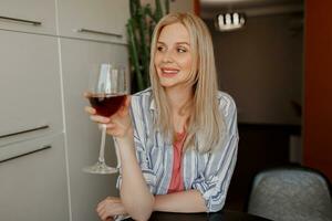 blond Frau halten Glas von rot Wein auf ihr besitzen Küche. foto