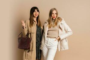 zwei elegant Frauen im stilvoll Winter Outfit posieren Über Beige Hintergrund. tragen Mantel und Jacke. Einkaufen und Mode Konzept. foto